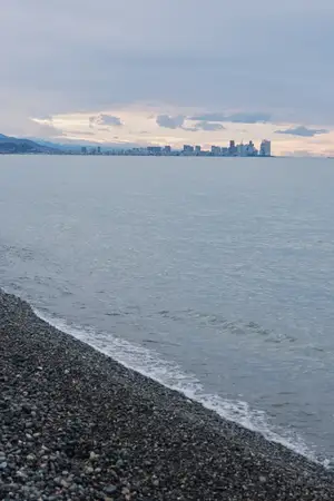 Тихая сцена на берегу: галечный пляж, спокойная вода и силуэт городского горизонта на горизонте создают безмятежный прибрежный вид в сумерках
