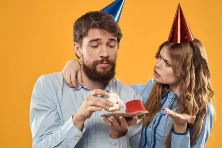 Мужчина и женщина веселятся на праздновании дня рождения на жёлтом фоне, на них праздничные колпаки