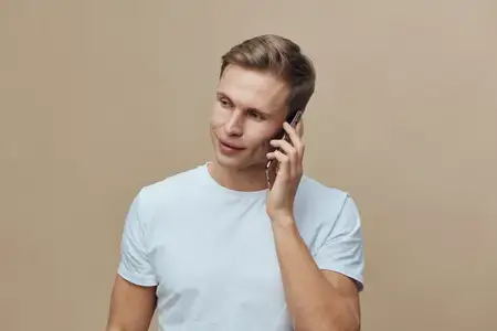 Улыбающийся мужчина пользуется мобильным телефоном, на нем повседневная белая футболка, помещение с однотонной бежевой стеной, естественное освещение, расслабленное настроение