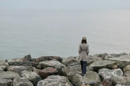 Женщина в нейтральной одежде стоит на камнях у моря и смотрит на воду