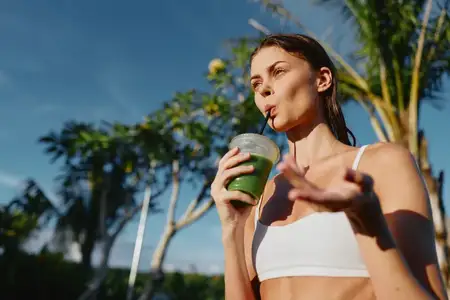 Спортивная женщина в белом топе-кропе пьёт зелёный сок на улице в солнечный день рядом с пальмами, воплощая здоровье и активный образ жизни с освежающим полезным напитком