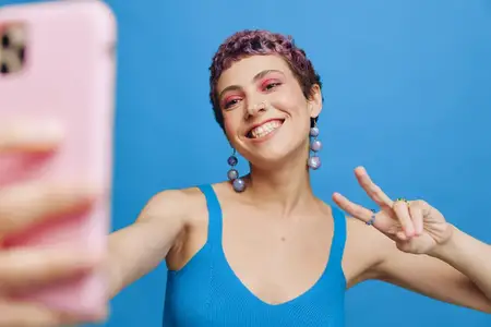 Молодая спортивная женщина, модный блогер, фотографирует себя на телефон в синей спортивной одежде, улыбается и показывает язык на однотонном синем фоне
