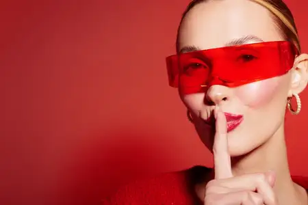 Крупный план молодой женщины в красных солнцезащитных очках и красном свитере, прикладывающей палец к губам в жесте «тише»