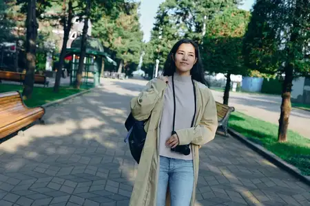 Уличная сцена: стильная женщина в парке, в тренчкоте и джинсах, с рюкзаком через плечо
