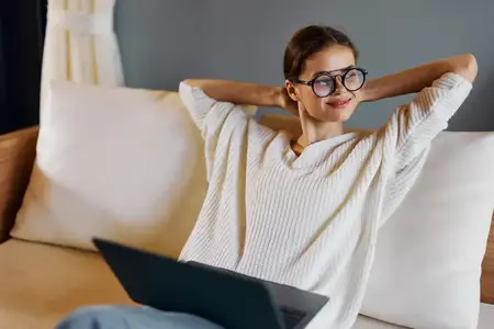 Улыбающаяся женщина работает за ноутбуком, уютно устроившись на диване в своем современном домашнем офисе