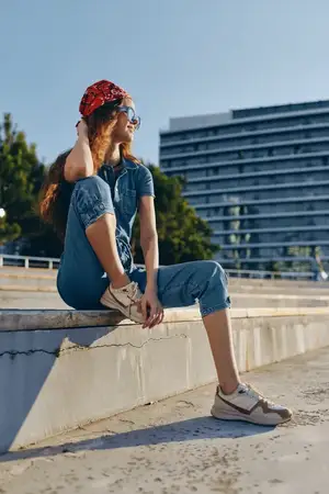 Женщина в стиле casual, одетая в современный бохо-вестерн деним стритстайл, расслабленно сидит на улице на бетонных ступенях, в красной бандане и солнцезащитных очках, залитая теплым солнечным…