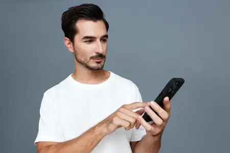 Молодой человек, увлеченно использующий свой смартфон, одетый в простую белую футболку, демонстрирует задумчивый вид