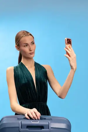Женщина-путешественница в темно-зеленом платье делает селфи на смартфон рядом с чемоданом