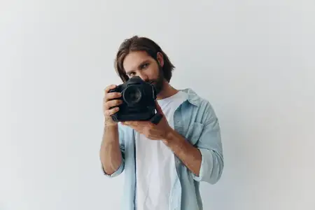 Мужчина-хипстер-фотограф в студии на фоне белого фона держит профессиональную камеру и настраивает её перед съёмкой