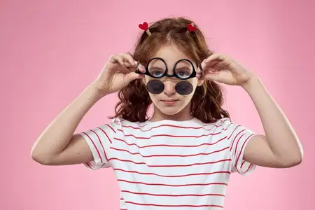 Маленькая девочка в модных очках и с зажимами для волос, её эмоции на розовом фоне с сердечками