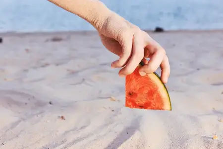 Крупный план руки, берущей кусок арбуза из песка на солнечном пляже, что вызывает летнее настроение