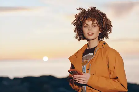 Женщина с вьющимися волосами в оранжевой куртке держит смартфон на закате, на открытом воздухе у побережья, портрет в стиле жизни, золотой вечерний свет, расслабленное выражение для путешествий…