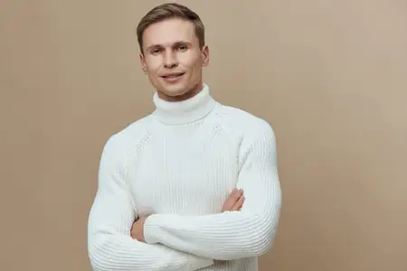 Улыбающийся молодой человек со светлой кожей и короткими каштановыми волосами, одетый в уютный белый вязаный свитер с высоким воротником, стоит со скрещенными руками и расслабленным выражением лица