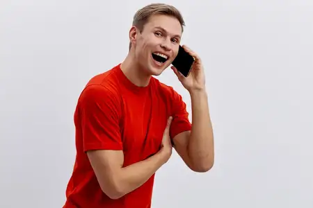 Счастливый молодой человек в яркой красной футболке ведет оживленный телефонный разговор на нейтральном белом фоне, демонстрируя позитивные эмоции и современный стиль общения