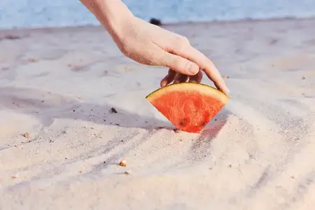 Рука аккуратно кладёт ломтик арбуза на светлый песок на пляже, что символизирует лето, отдых и свежесть