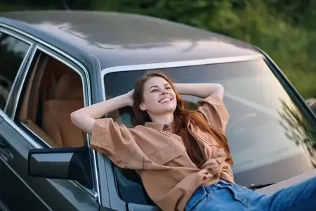 Веселая молодая женщина в повседневной одежде наслаждается отдыхом на винтажном автомобиле в окружении пышной зелени, излучая беззаботное летнее настроение
