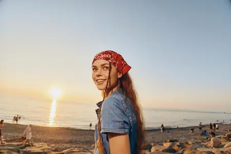 Счастливая женщина в повседневном образе, одетая в современный бохо-вестерн деним и красную бандану, наслаждается закатом на каменистом пляже