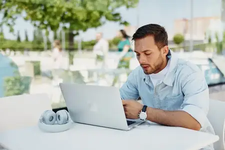 Сосредоточенный молодой человек работает за ноутбуком, сидя за уличным столиком в солнечном парке