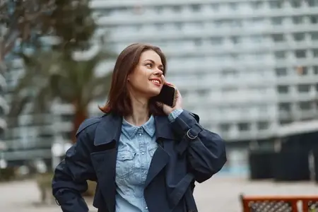 Женщина в джинсовой рубашке и темно-синей куртке стоит в городской обстановке на открытом воздухе, разговаривая по мобильному телефону на фоне города