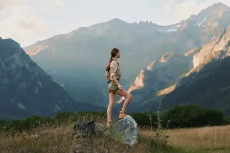 На ней майка, шорты, походные ботинки и небольшой рюкзак; она стоит, поставив одну ногу на камень, а долина простирается позади нее, излучая спокойную решимость