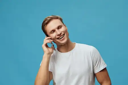 Улыбающийся молодой мужчина-европеец в повседневной белой футболке разговаривает по мобильному телефону на синем студийном фоне