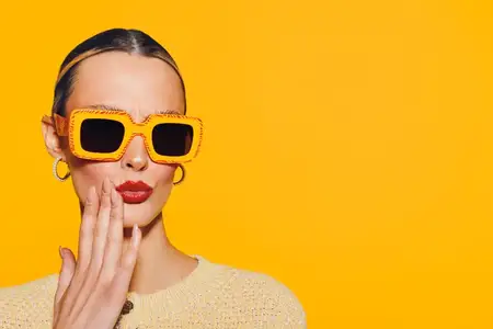 Уверенная женщина в больших оранжевых солнцезащитных очках и с красной помадой прикасается к лицу на ярко-желтом фоне