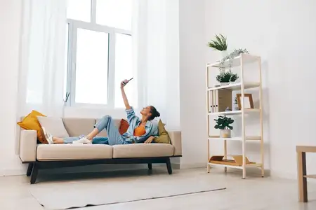 Весёлая женщина держит смартфон и улыбается, наслаждаясь досугом на уютном диване дома
