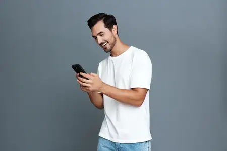 Радостный улыбающийся мужчина взаимодействует со смартфоном, одетый в повседневную белую футболку и синие джинсы, на сером фоне, воплощая современный образ жизни и вовлеченность