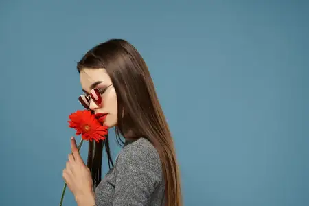 Красивая брюнетка в солнцезащитных очках с красным цветком у лица, косметика на синем фоне