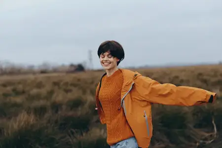 Молодой человек улыбается на открытом воздухе, одет в оранжевую куртку в поле, окружен природой: лугами и облачным небом, мягкий студийный дневной свет, образ жизни для повседневных путешествий…