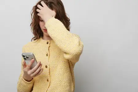Стрессующая молодая женщина в желтом вязаном свитере держит смартфон и касается лба с закрытыми глазами, демонстрируя эмоциональную усталость и тревогу