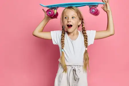 Улыбающаяся маленькая девочка с двумя длинными косичками держит голубой скейтборд над головой на розовом фоне