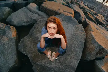 Молодая женщина в стиле лайфстайл, одетая в современный бохо-вестерн деним, сидит на фактурных камнях у берега, снятая с теплыми кинематографическими цветовыми оттенками, усиливающими естественную…
