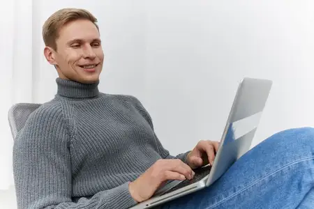 Улыбающийся мужчина работает за ноутбуком в сером свитере и синих джинсах, сидя расслабленно у белой стены, используя современные технологии для удаленной работы и образа жизни цифрового кочевника…
