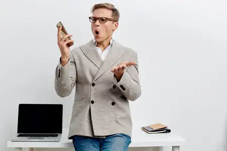 Удивлённый мужчина разговаривает по мобильному телефону, одетый в серый блейзер и синие джинсы, стоит рядом с белым столом, на котором лежат ноутбук и блокнот, на чистом белом фоне