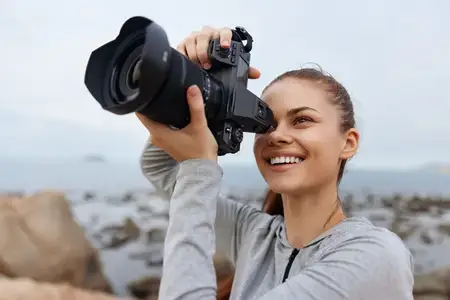 Счастливая молодая женщина-фотограф в серой одежде радостно фотографирует на пляже, в окружении камней и воды, воплощая творчество и дух приключений