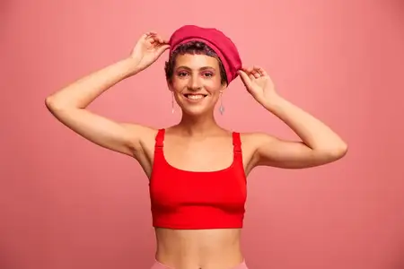 Молодая спортивная женщина с короткой стрижкой и фиолетовыми волосами в красном топе и розовой шапке, со спортивной фигурой, улыбается и гримасничает, показывая знаки руками, и смотрит в камеру на…
