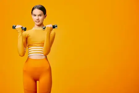 Спортивная женщина в подходящем оранжевом костюме выполняет упражнение для верхней части тела с гантелями, подчеркивая целеустремленность, осанку и энергию в яркой студийной обстановке