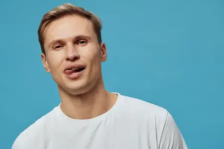 Портрет юмористического молодого человека со светлой кожей, короткими каштановыми волосами, одетого в простую белую футболку, с игривым, глупым выражением лица на фоне синего фона