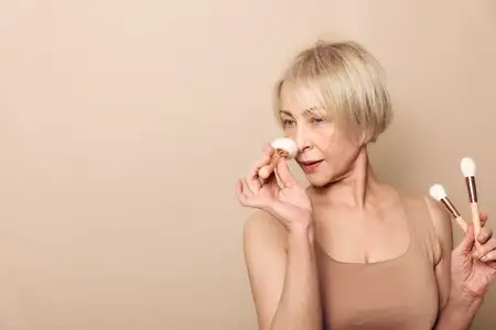 Светловолосая пожилая женщина, держа кисти для макияжа, аккуратно наносит пудру на лицо