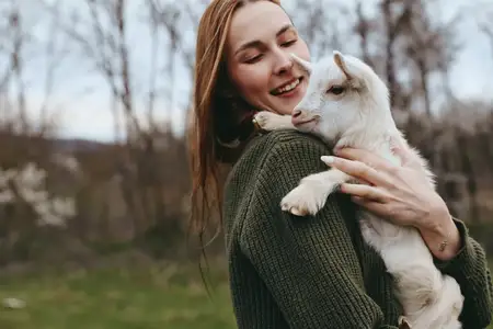 Молодая женщина в зеленом свитере с любовью обнимает белого козленка в естественной природной среде с голыми деревьями на заднем плане осенним днем