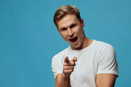 Возбужденный молодой мужчина-европеец в белой футболке указывает пальцем на камеру с открытым ртом и энергичным выражением лица, изолирован на простом синем фоне, студийный портрет