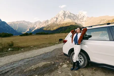 Женщина стоит рядом с припаркованной машиной на фоне суровых гор и открытого пейзажа на грунтовой дороге, воплощая путешествие и отдых на природе в повседневном стиле, свободу и исследование природы