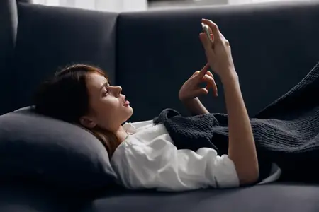 Женщина, укрытая одеялом на диване, смотрит на экран мобильного телефона