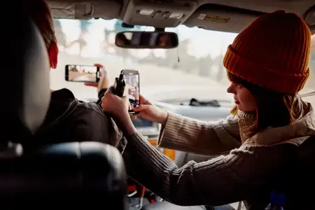 Женщина в шляпе сидит в салоне автомобиля с телефоном в руках, вокруг природа
