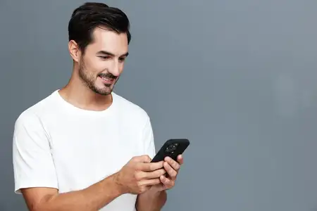 Радостный улыбающийся мужчина уверенно стоит, проверяя свой смартфон, одетый в повседневную белую футболку на фоне светло-серого фона