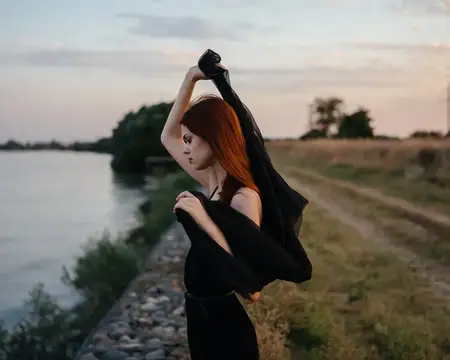 Женщина в чёрном платье с тканью возле лица отдыхает на природе у реки