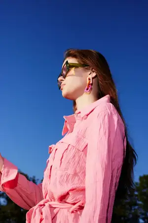 Модная молодая женщина в розовом платье-рубашке с большими солнцезащитными очками стоит на фоне ярко-голубого неба, излучая уверенность и летнюю элегантность