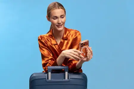 Улыбающаяся путешественница в оранжевой рубашке пользуется смартфоном на фоне темно-синего багажа и голубого фона