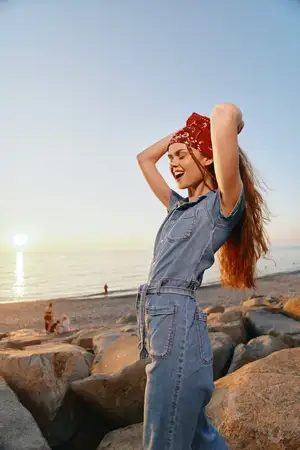 Женщина, ведущая активный образ жизни, одетая в современный бохо-вестерн деним в стиле стрит-стайл, счастливо позирует на каменистом пляже в золотой час